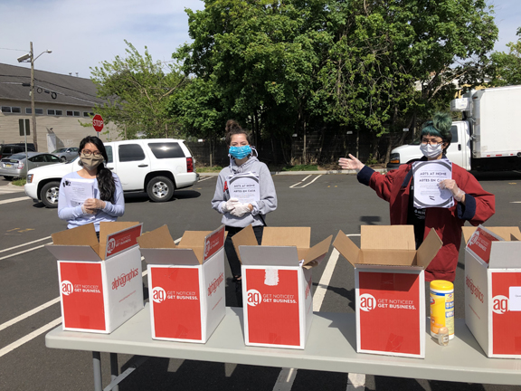 Three women in masks stand behind a gray table of red and white boxes full of packets to hand out