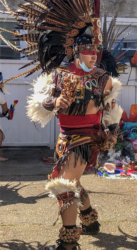 Photo of a Native American man in Aztec-inspired garb dancing. He has long light and dark feathers on his large beaded headdress and short white feathers on his arms. On his chest and wrists is beautiful, multicolored beadwork.