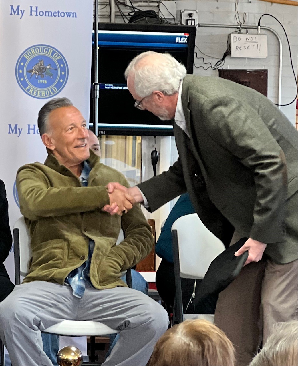 Photo of Bruce Springsteen, sitting, shaking the hand of another gentleman, who is standing and facing away from the camera