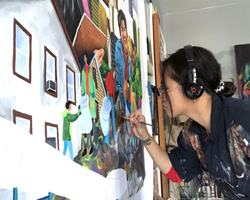 Photo of a young woman with dark hair painting. She is wearing gold wire glasses and black headphones. Her navy blue collared shirt is splattered with paint. 