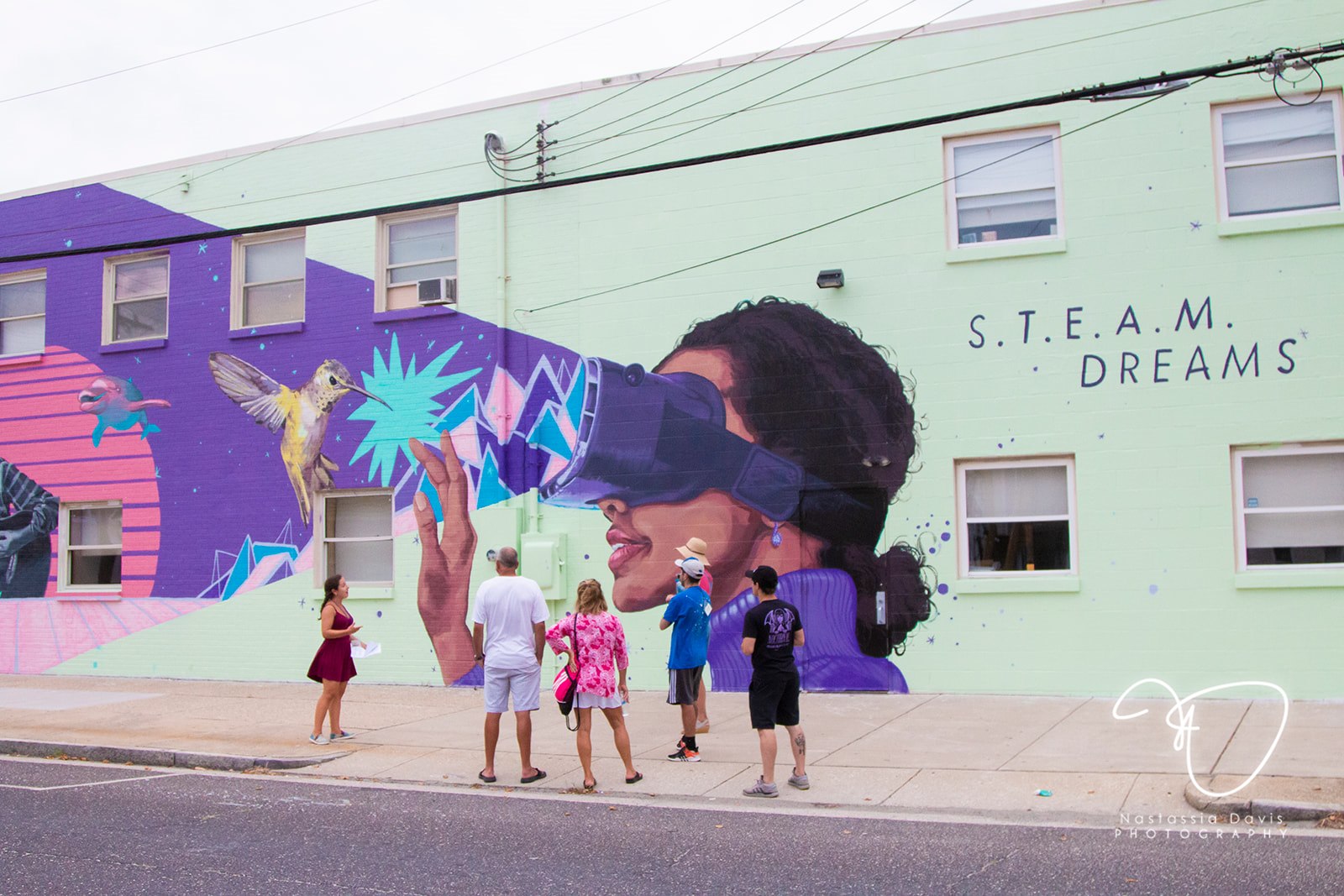 Photo of a mural by LNY titled S.T.E.A.M. DREAMS. The mural features the face of Black female-presenting individual in a purple shirt. She is wearing navy goggles that seem to be showing her a grey, white and yellow hummingbird, as well as a dolphin and various shapes that are blue, pink and turquoise. There are six individuals of varying heights and ages standing in front of the mural.